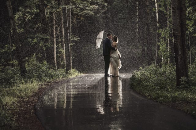 Bride and groom kissing in the rain on a path between trees, under white umbrella